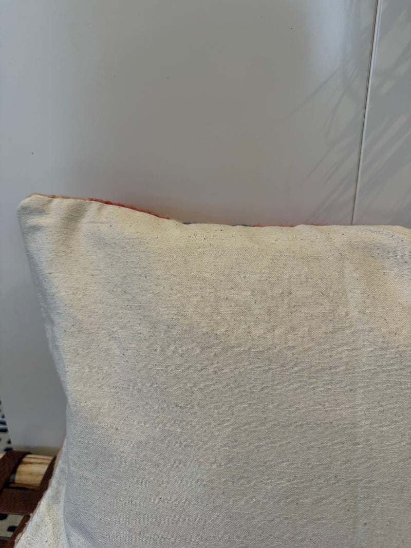 A close-up of a square throw pillow with a textured cream linen cover, showing a hint of red piping along the top edge.