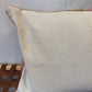 A square off-white linen throw pillow with a subtle multicolor embroidered border, resting on a brown woven wooden bench against a light gray wall.