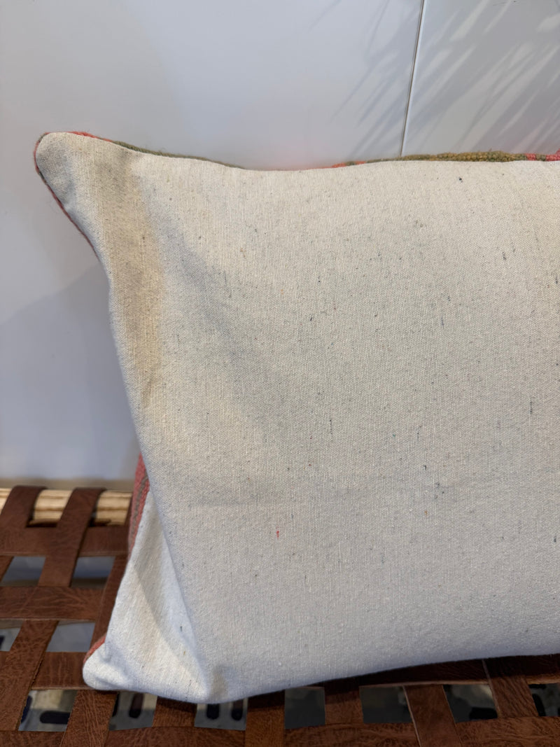 A square off-white linen throw pillow with a subtle multicolor embroidered border, resting on a brown woven wooden bench against a light gray wall.