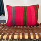 A square throw pillow with a vibrant red base and vertical green and gray stripes, resting on a rustic woven wooden bench.