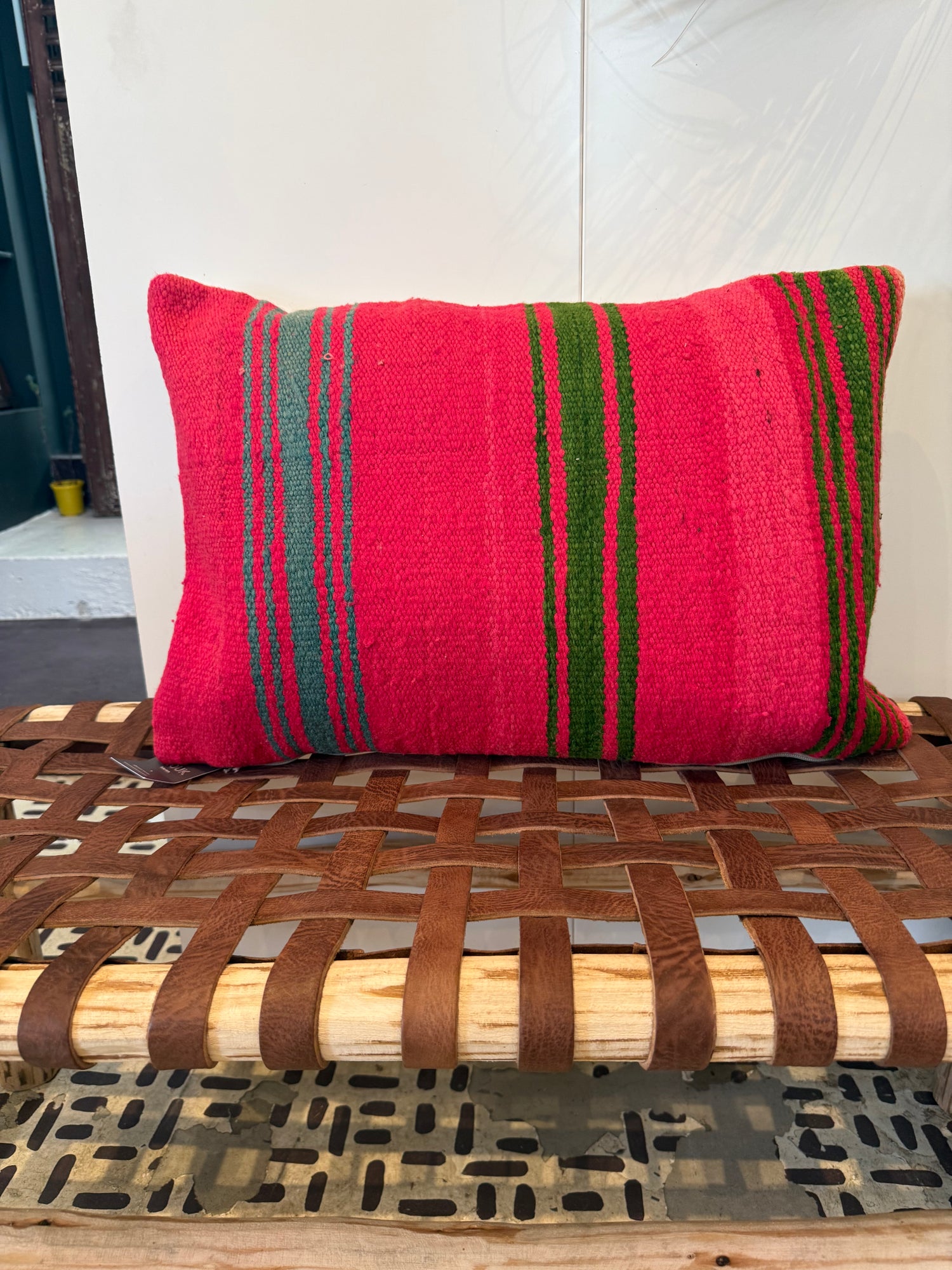 A square throw pillow with a vibrant red base and vertical green and gray stripes, resting on a rustic woven wooden bench.