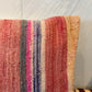 A close-up of a woven throw pillow cover featuring vertical stripes in pink, red, gray, blue, and beige, set against a light gray wall with leaf shadows.