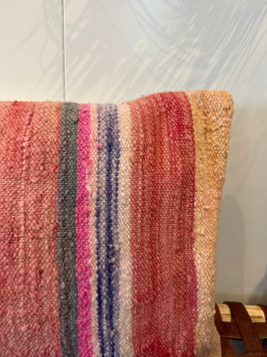 A close-up of a woven throw pillow cover featuring vertical stripes in pink, red, gray, blue, and beige, set against a light gray wall with leaf shadows.