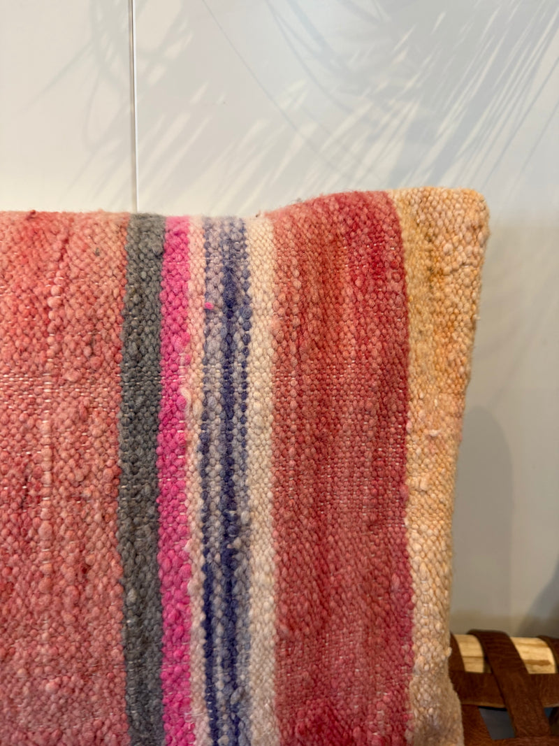 A close-up of a woven throw pillow cover featuring vertical stripes in pink, red, gray, blue, and beige, set against a light gray wall with leaf shadows.
