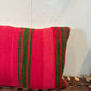 A square woven throw pillow in vibrant pink with vertical green stripes, resting on a brown leather-wrapped wooden bench against a white wall.