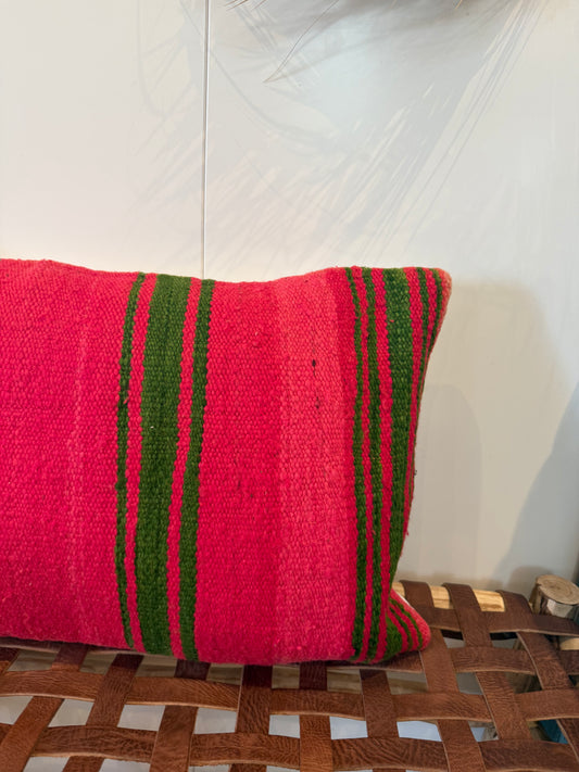 A square woven throw pillow in vibrant pink with vertical green stripes, resting on a brown leather-wrapped wooden bench against a white wall.