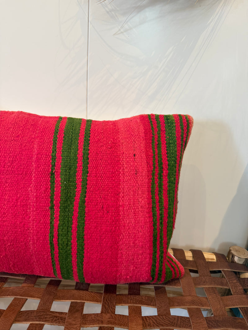 A square woven throw pillow in vibrant pink with vertical green stripes, resting on a brown leather-wrapped wooden bench against a white wall.