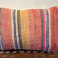 A rectangular woven throw pillow with vertical stripes in pink, orange, yellow, blue, and cream, resting on a brown woven bench against a white wall.