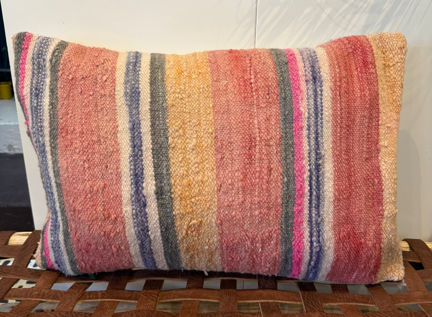 A rectangular woven throw pillow with vertical stripes in pink, orange, yellow, blue, and cream, resting on a brown woven bench against a white wall.