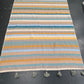 A rectangular woven rug with horizontal stripes in mustard yellow, teal, light blue, beige, and white, finished with gray tassels on the top and bottom edges, laid flat on a dark gray floor.