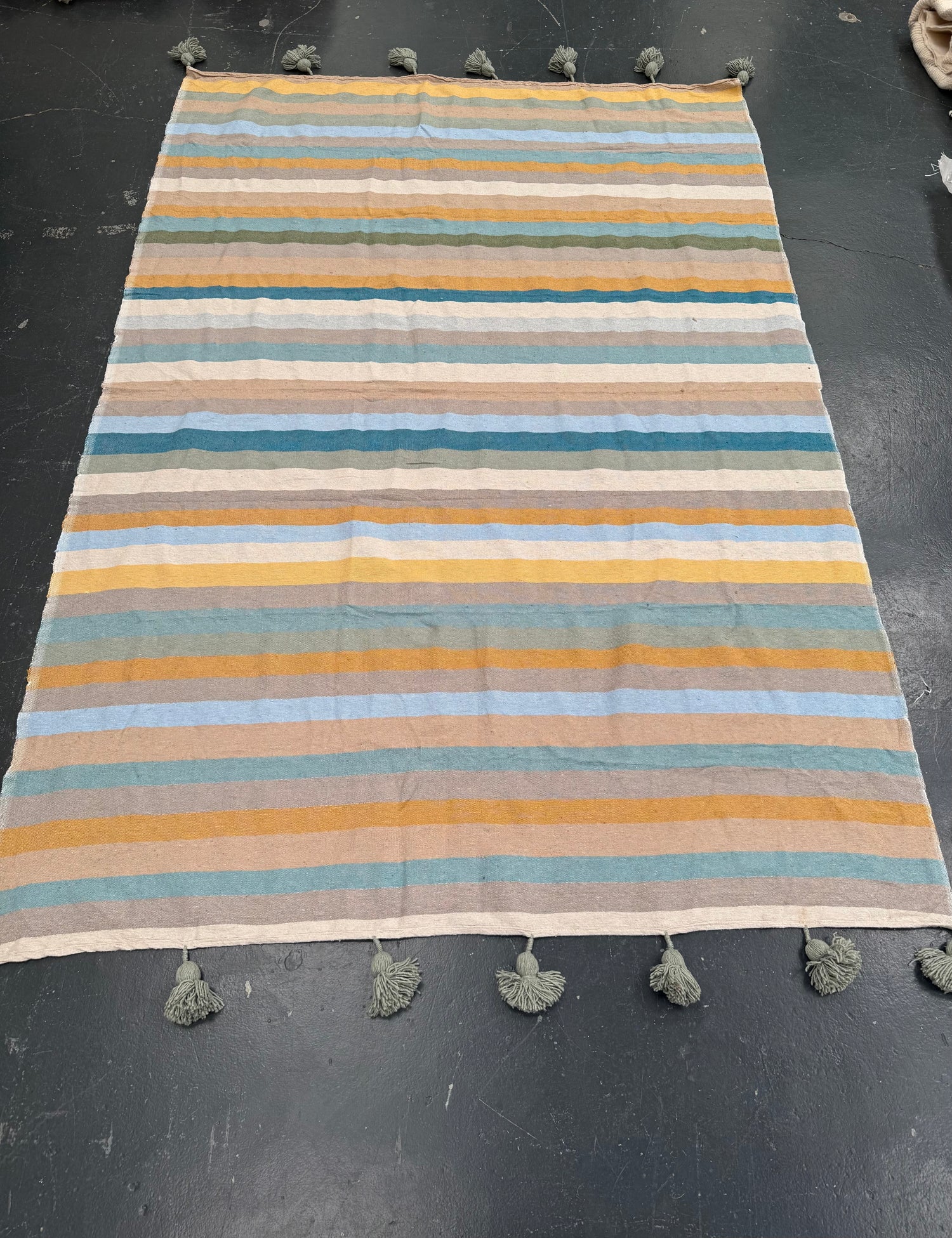 A rectangular woven rug with horizontal stripes in mustard yellow, teal, light blue, beige, and white, finished with gray tassels on the top and bottom edges, laid flat on a dark gray floor.