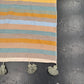A close-up of a striped textile runner or rug with horizontal bands in cream, mustard yellow, light blue, sage green, and taupe, finished with three knotted tassels on the bottom edge, laid on a dark textured surface.