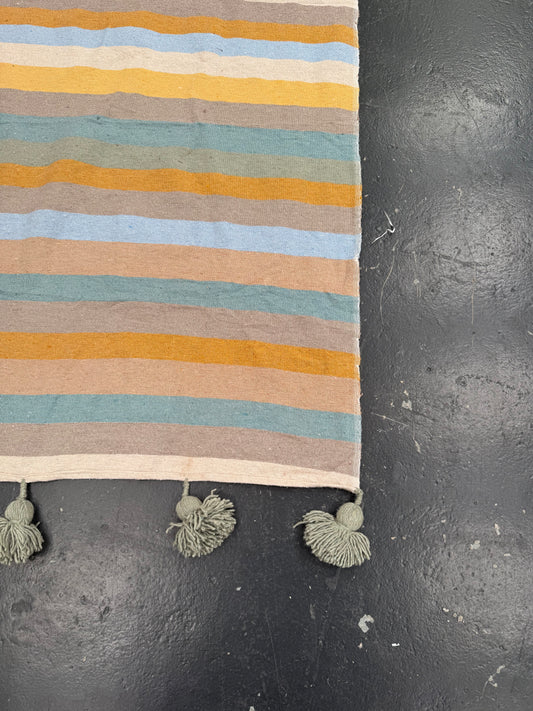 A close-up of a striped textile runner or rug with horizontal bands in cream, mustard yellow, light blue, sage green, and taupe, finished with three knotted tassels on the bottom edge, laid on a dark textured surface.
