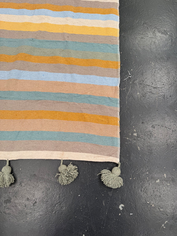 A close-up of a striped textile runner or rug with horizontal bands in cream, mustard yellow, light blue, sage green, and taupe, finished with three knotted tassels on the bottom edge, laid on a dark textured surface.