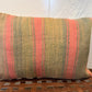A rectangular woven throw pillow with vertical stripes in muted olive green, dusty pink, and tan, resting on a brown woven leather bench against a white wall.