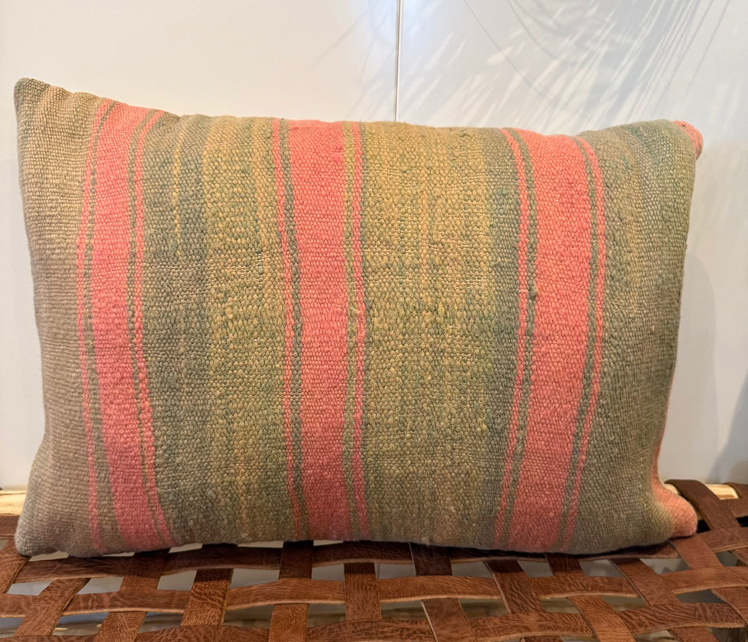 A rectangular woven throw pillow with vertical stripes in muted olive green, dusty pink, and tan, resting on a brown woven leather bench against a white wall.