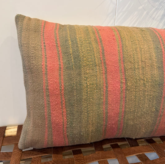 A rectangular woven throw pillow with vertical stripes in muted olive green, terracotta pink, and tan, resting on a wooden bench against a white wall.