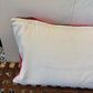 A square white pillow with a red and green patterned fabric trim, resting on a brown woven wooden bench against a white wall.