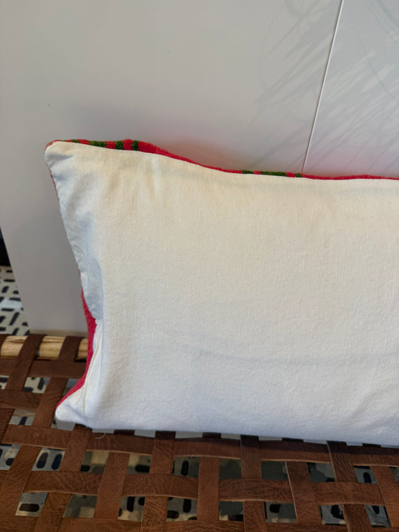 A square white pillow with a red and green patterned fabric trim, resting on a brown woven wooden bench against a white wall.