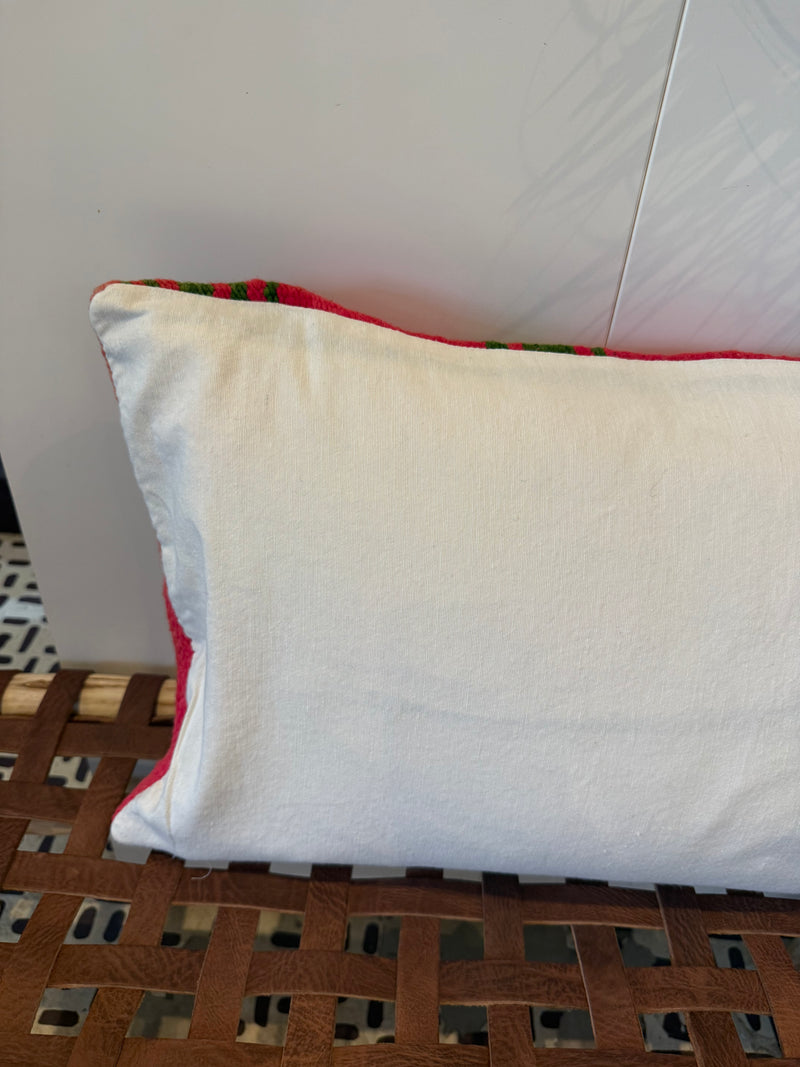 A square white pillow with a red and green patterned fabric trim, resting on a brown woven wooden bench against a white wall.